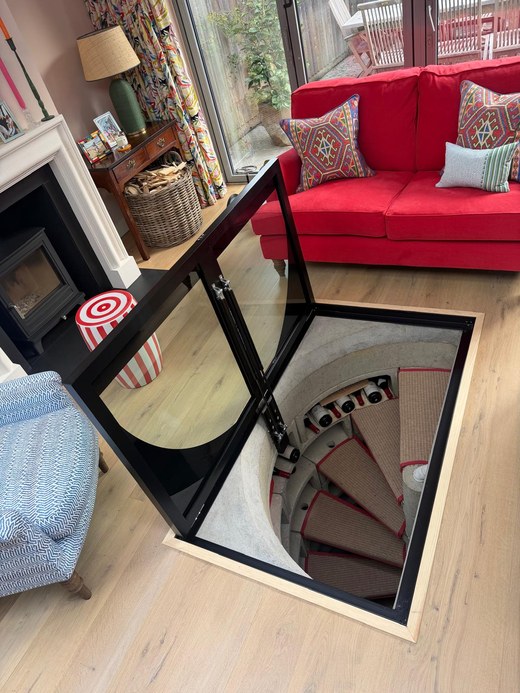 Hinged glass cellar door standing open on gas struts above a spiral staircase in a London living room