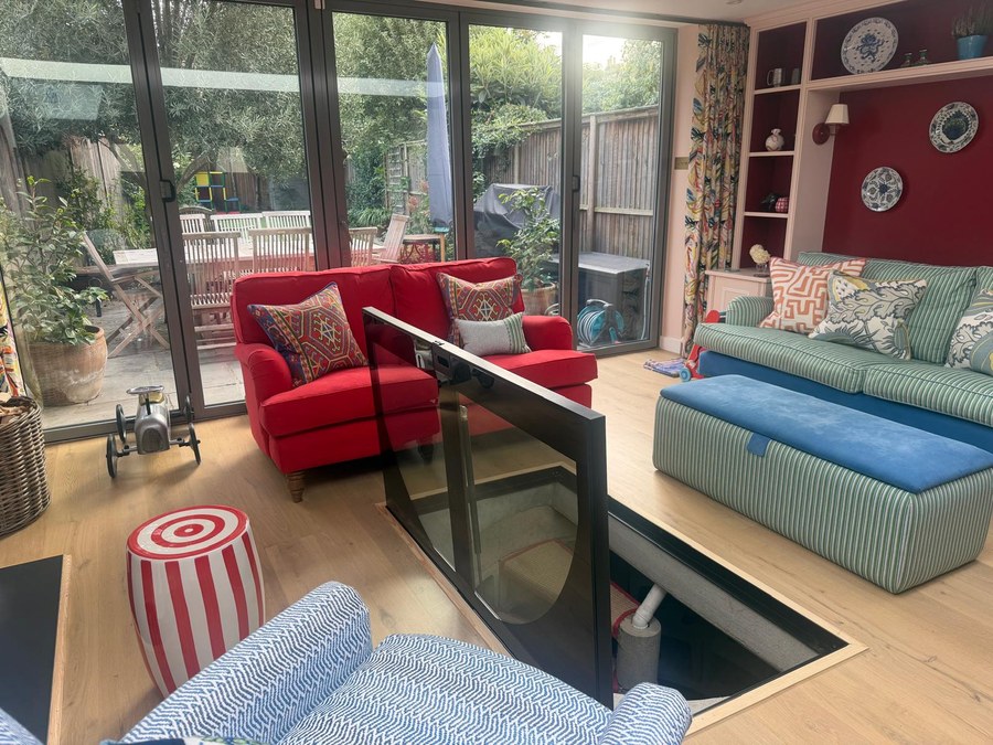 Hinged glass floor door held open on gas struts in a living room with garden doors behind