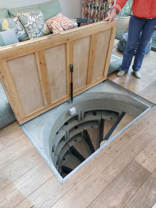 Timber plywood trapdoor propped open over concrete cellar stairs in a London living room