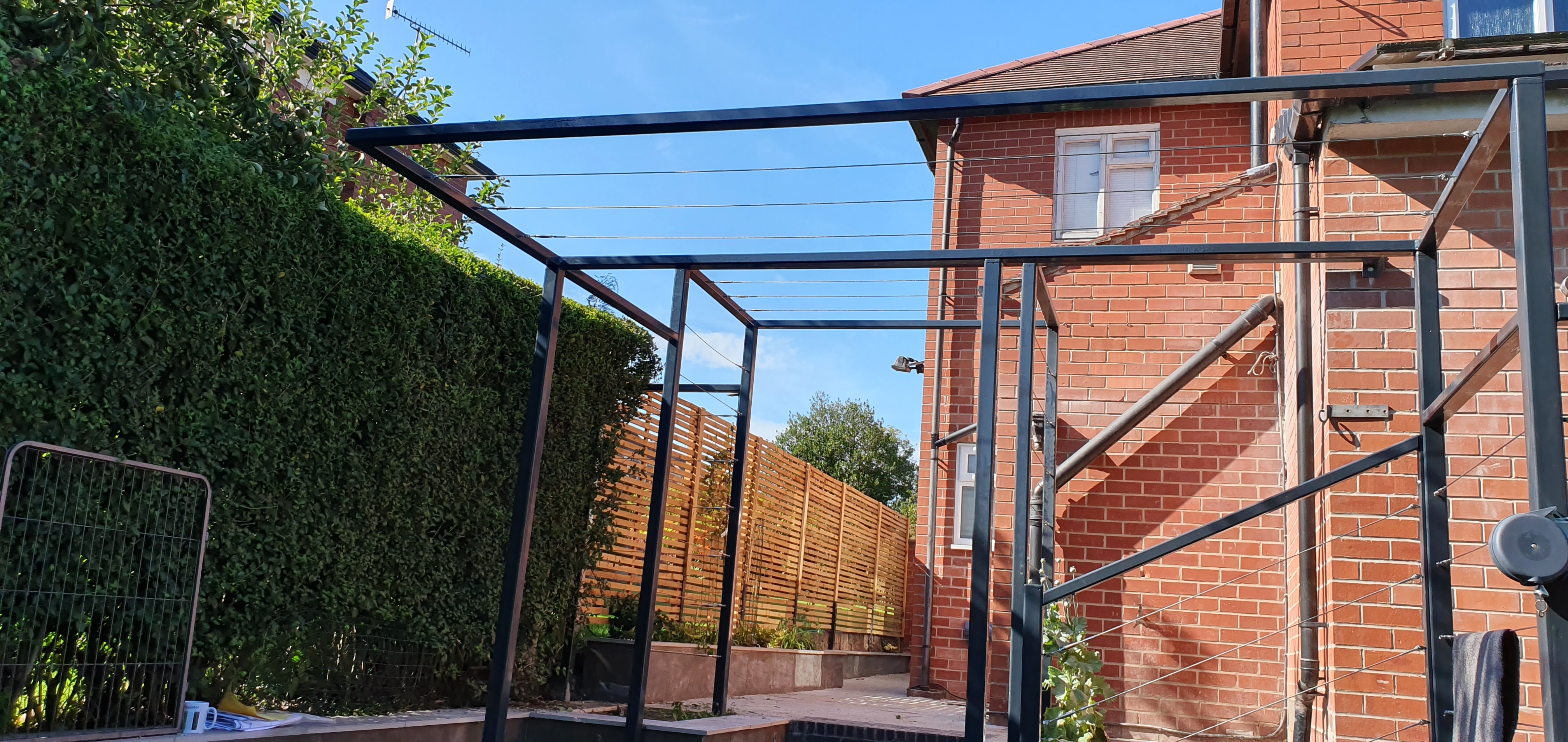 Powder coated steel pergola with tensioned stainless wires across the top against a brick house