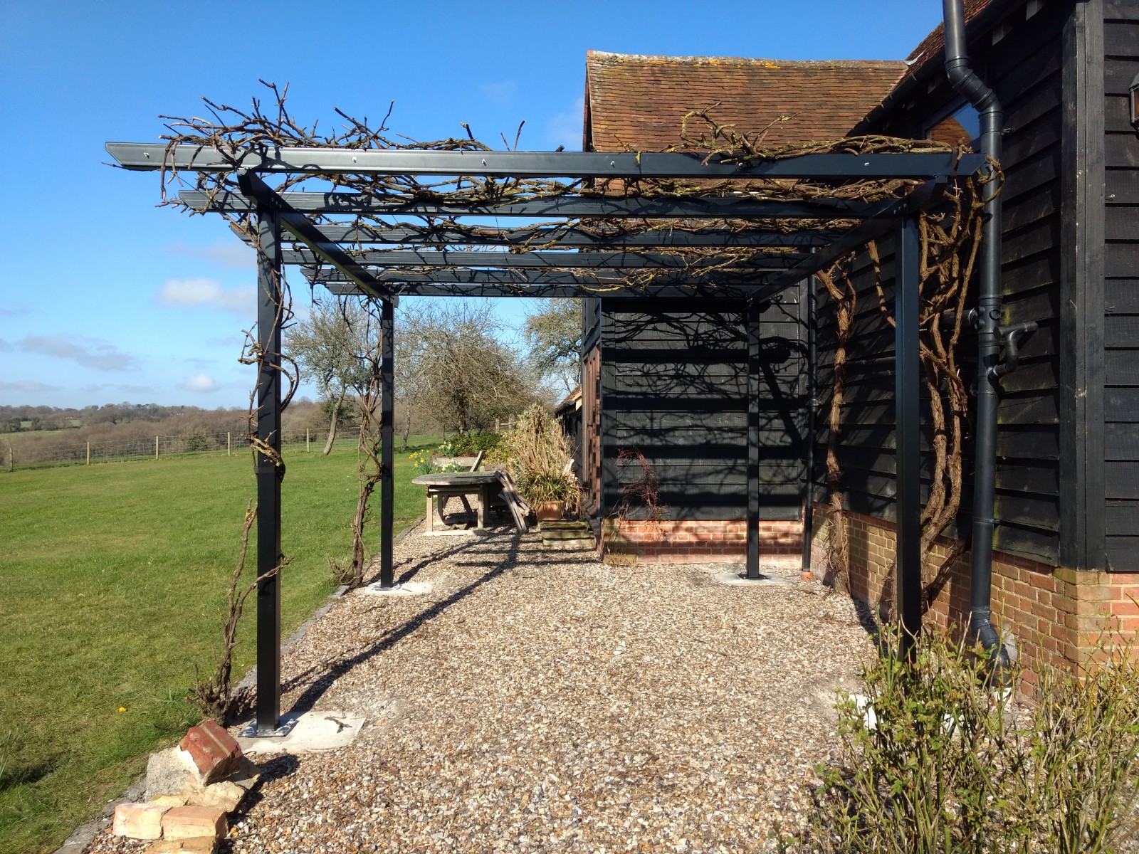 Long black powder coated steel pergola with close spaced rafters over a gravel terrace