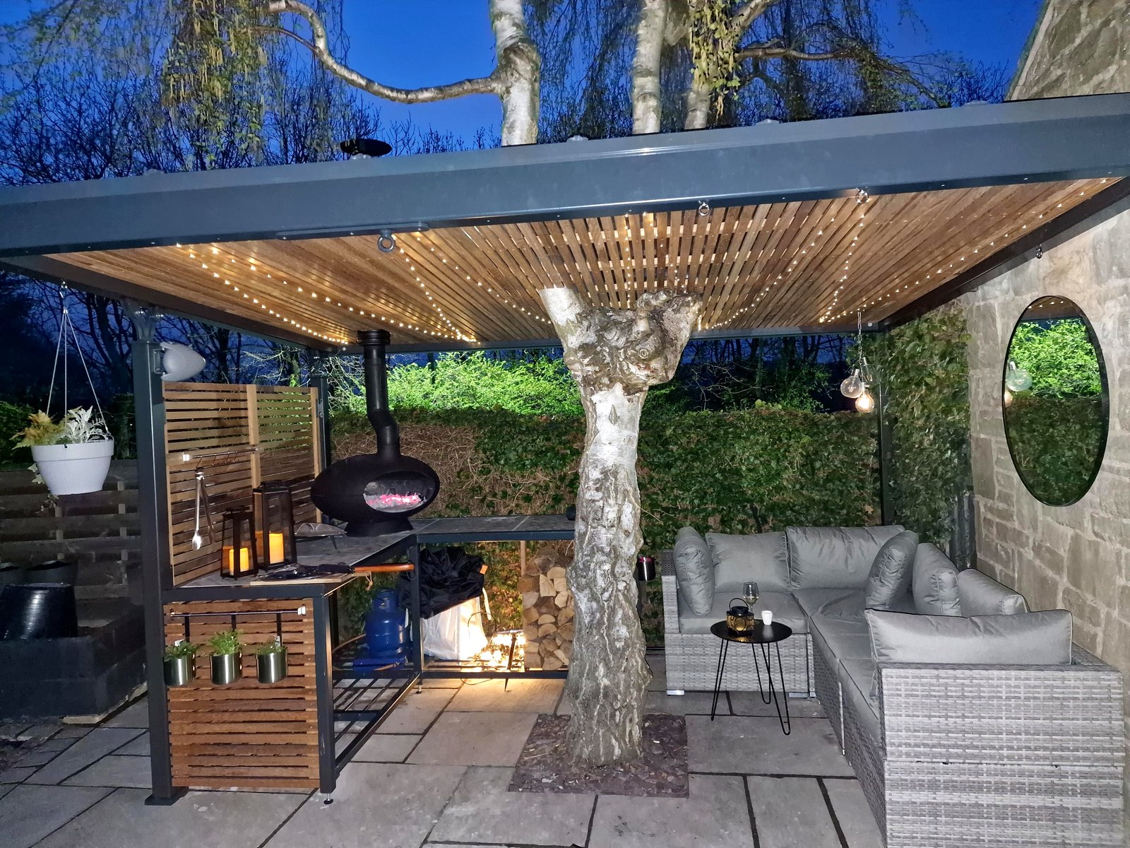 Steel pergola with timber-slatted roof and fairy lights at night, around a tree, Sheffield