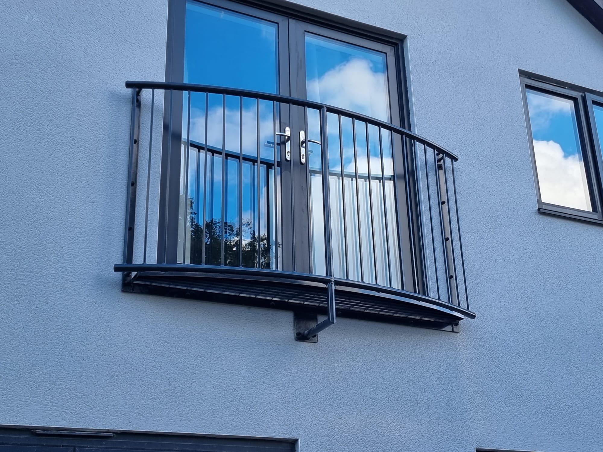 Curved step-out Juliet balcony in black powder coated steel on a rendered house, showing the shallow platform and support bracket